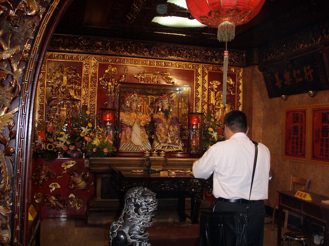 A white-shirted man with a briefcase worships at the temple made for Matsu. Next to him is a playful lion statue. The temple sits in the Dajia District of Taichung, Taiwan.